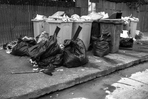 A row of large outdoor rubbish bins situated on a paved surface, with one beige bin and another darker grey bin, both filled with various waste materials and lids partially open. Several black and transparent plastic rubbish bags, some bulging and tied at the top, are piled in front of and beside the bins, containing discarded items. The waste appears to include paper, cardboard, and miscellaneous debris. The bins are positioned near a wooden fence, with a grassy area visible beyond the fence in the background. The scene is illuminated by natural daylight, casting shadows on the pavement, and the area appears to be in a residential or commercial setting in Finchley, London. This image relates to rubbish removal services offered by Commercial Waste Removal Finchley, showcasing typical waste accumulation that requires professional collection and disposal.