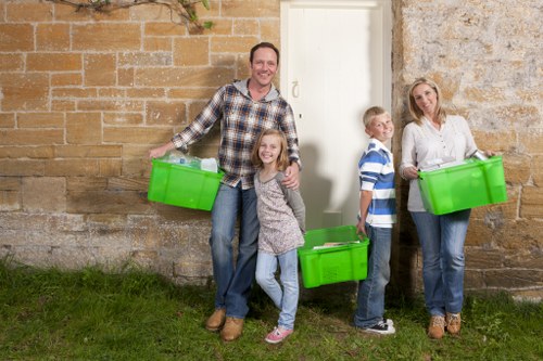 A family of four standing outdoors in front of a historic stone wall with a white door, each holding bright green plastic storage boxes. The father, wearing a plaid shirt and jeans, is positioned on the left with a box in his right hand, smiling at the camera. Next to him, a young girl with long hair and casual clothing is also holding a green container, standing close and smiling. To her right, a boy in a striped shirt and jeans holds a similar green box, looking towards the camera. On the far right, a woman with blonde hair, dressed in a white blouse and jeans, holds another green container, also looking at the camera. The setting appears to be a residential or rural area with a grassy foreground and weathered stone wall background, likely within the Finchley area, suggesting a rubbish or waste collection context. The brightly coloured containers contrast with the neutral tones of the wall and ambient natural light highlights the scene, indicative of an outdoor waste management activity aligns with services provided by Commercial Waste Removal Finchley.